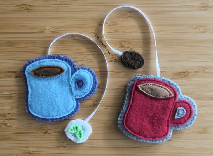 Two felt bookmarks are laid flat on a wood surface, photographed from above. They look like two mugs of hot beverages, one mug red and one blue. Connected to them with ribbon are two tags; one a teabag label and the other a coffee bean.