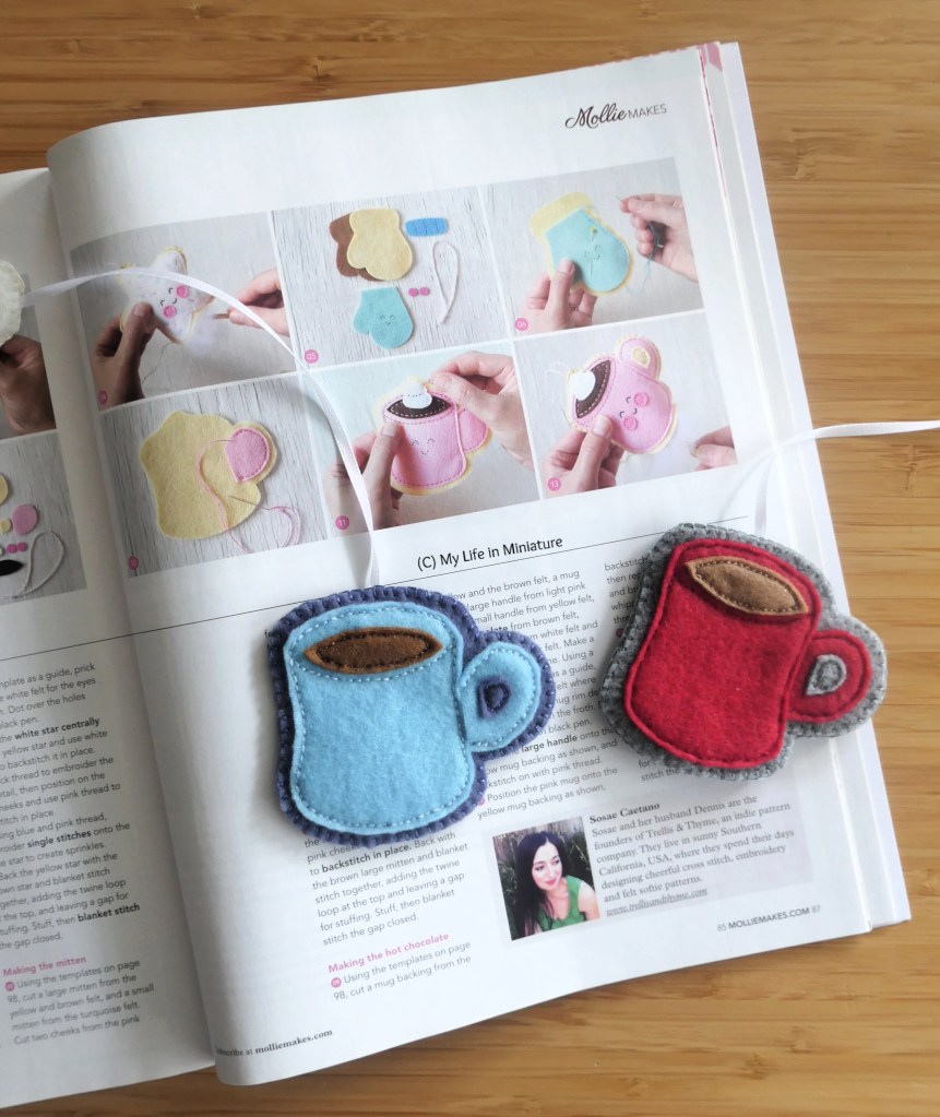 The two mug bookmarks are laid out on an open magazine. The magazine is open to a page with images of the making process of a similar felt mug-with-a-hot-drink-inside. 