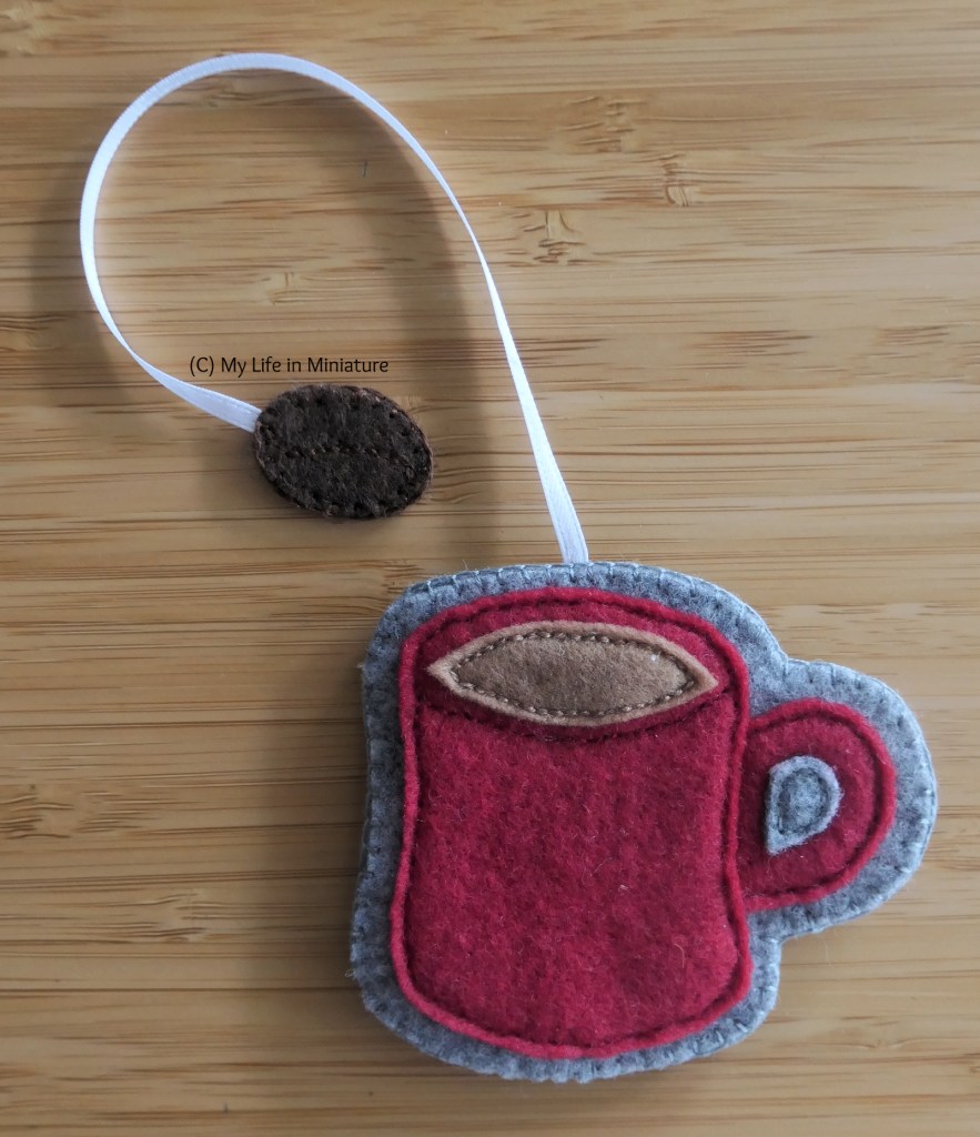 Closeup of the red mug. It is on a grey background, and has a lighter brown liquid in. Its ribbon is also white, and it has a dark brown coffee bean attached to the end.
