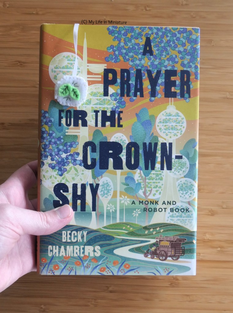 The author's white hand is holding 'A Prayer for the Crown-Shy' by Becky Chambers over a wood surface. Visible on the front cover of the book is the teabag square end of the pale blue bookmark. 