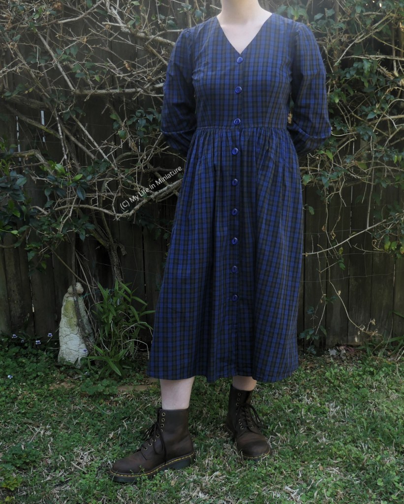 The author stands in front of a hedge, photographed from neck to the ground. She wears a navy blue V-necked checked dress with buttons down the front. She also has brown boots on, and her hands are behind her back.