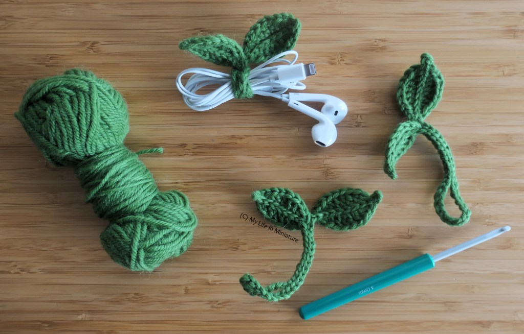 Several small objects are photographed from above on a wood surface. There is a ball of green yarn, a crochet hook, a pair of wired earbuds wrapped in green yarn and crocheted leaves, and two unwrapped crochet yarn/leaves. The yarn/leaves are two crocheted leaves, connected at one end to a small length of crochet chain with a loop at the end. They are small enough to loop around wires to hold them together. 