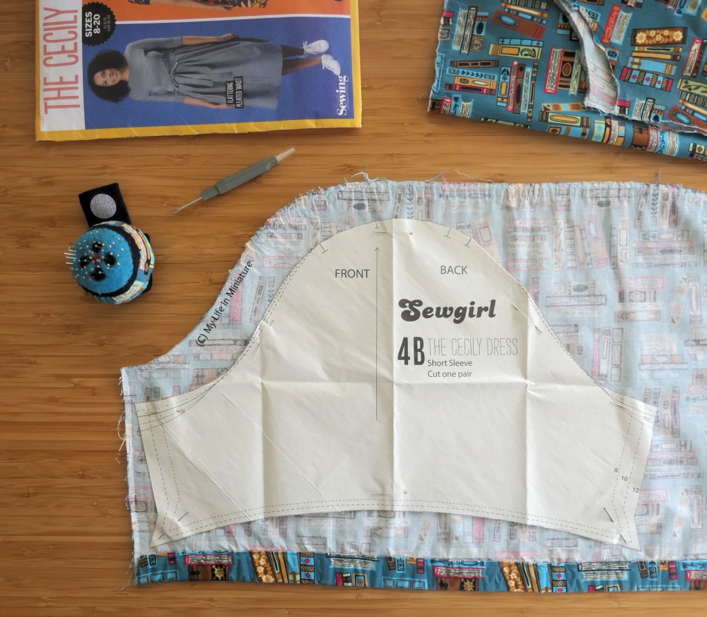 A sleeve is being cut out of a scrap of the book dress' fabric, photographed from overhead on a wood surface. The scrap looks like the old puffy sleeve of the dress, with a smaller sleeve pattern piece pinned to it. Around the fabric is scraps, a pincushion, an unpicker, and the pattern pouch for the dress. 