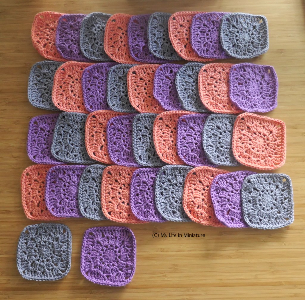 A number of crochet squares are laid flat on a wood surface, photographed from above. Most of them are in four rows of nine, with two extras at the bottom. They alternate each row in colour; peach, purple, and grey.