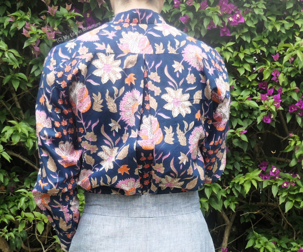 The author stands in front of a hedge, photographed from neck to hips. She faces away from the camera, so the back of the shirt is visible. The shirt is yoked, and has an inverted box pleat at the back to gather it.