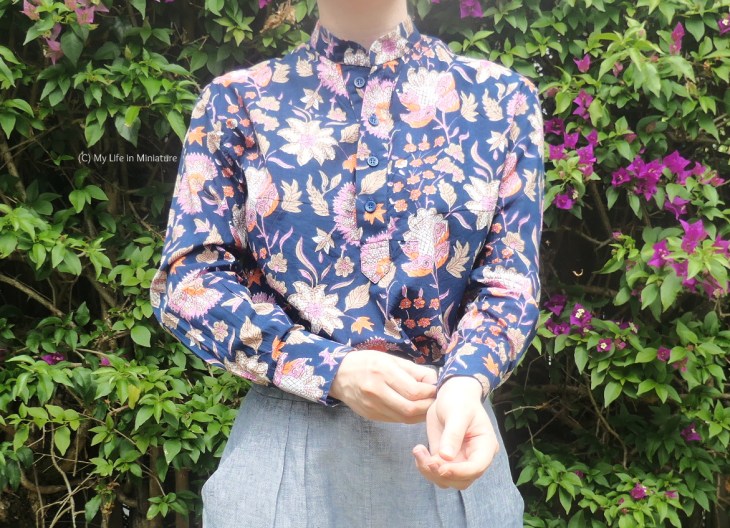 The author stands in front of a hedge, photographed fron neck to hips. She's wearing a floral button-down shirt tucked into pale grey pants. The shirt is navy with pink, cream, and peach florals, and is long-sleeved. The author is fiddling with one of the cuffs. 