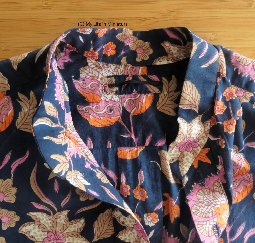 Close-up of the navy shirt, laid flat on a wood surface. The collar is open, so the inside yoke area is visible. There is a small rectangle sewn to the yoke, with a hanging loop of navy ribbon attached. 