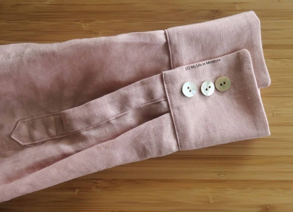 Close-up of the sleeve cuff and placket of a shirt in pale pink linen. The cuff has three small shell buttons resting on it. 