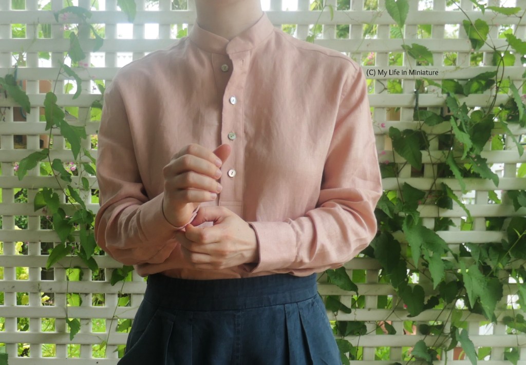 The author stands in front of some white lattice, with a vine growing through it. She's photographed from neck to hips, and is wearing a pale pink long-sleeved shirt with navy pants. She ahs her hands in front of her, and she's doing up the buttons on the cuffs of the sleeves. Her shirt is tucked into her pants.