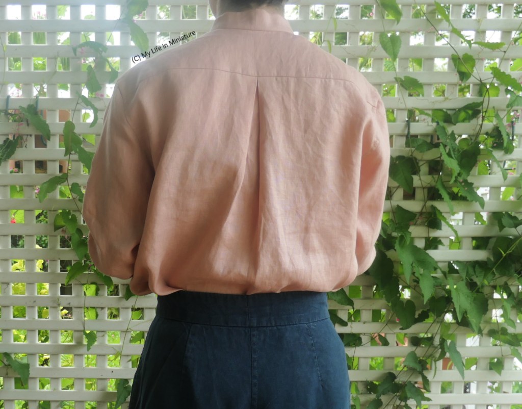 The author stands in front of a white lattice with a vine growing through it. She's photographed from neck to hips, and is wearing the same pale pink shirt and navy pants. Her back is to the camera, so the back of the shirt is visible. The shirt has a subtle box pleat at the centre of the yoke, and is tucked into the pants.
