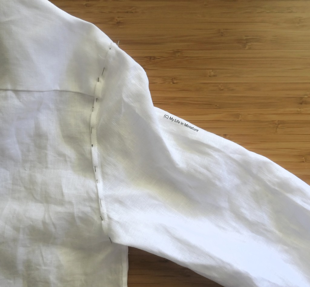 A white linen shirt is laid flat on a wood surface, photographed from above. The focus is on one of the armhole seams, where the sleeve attaches to the bodice. The seam allowance is folded down and pinned to the bodice. 