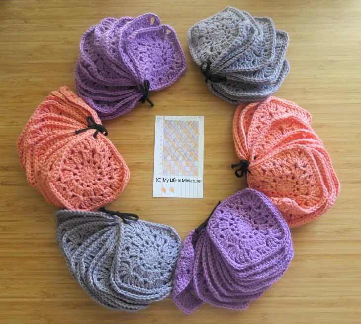 Six piles of crochet granny squares are arranged in a circle around a small notecard on a wood surface, photographed from above. The card tracks the progression of a blanket. The squares are peach, purple, and grey. 