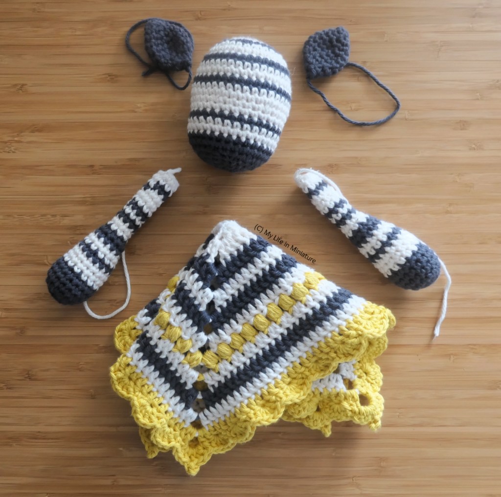 A disassembled crochet toy is laid flat on a wood surface. It has two ears beside its head, as well as two arms and a blanket folded into a square-ish shape below. All of the ends are loose. 