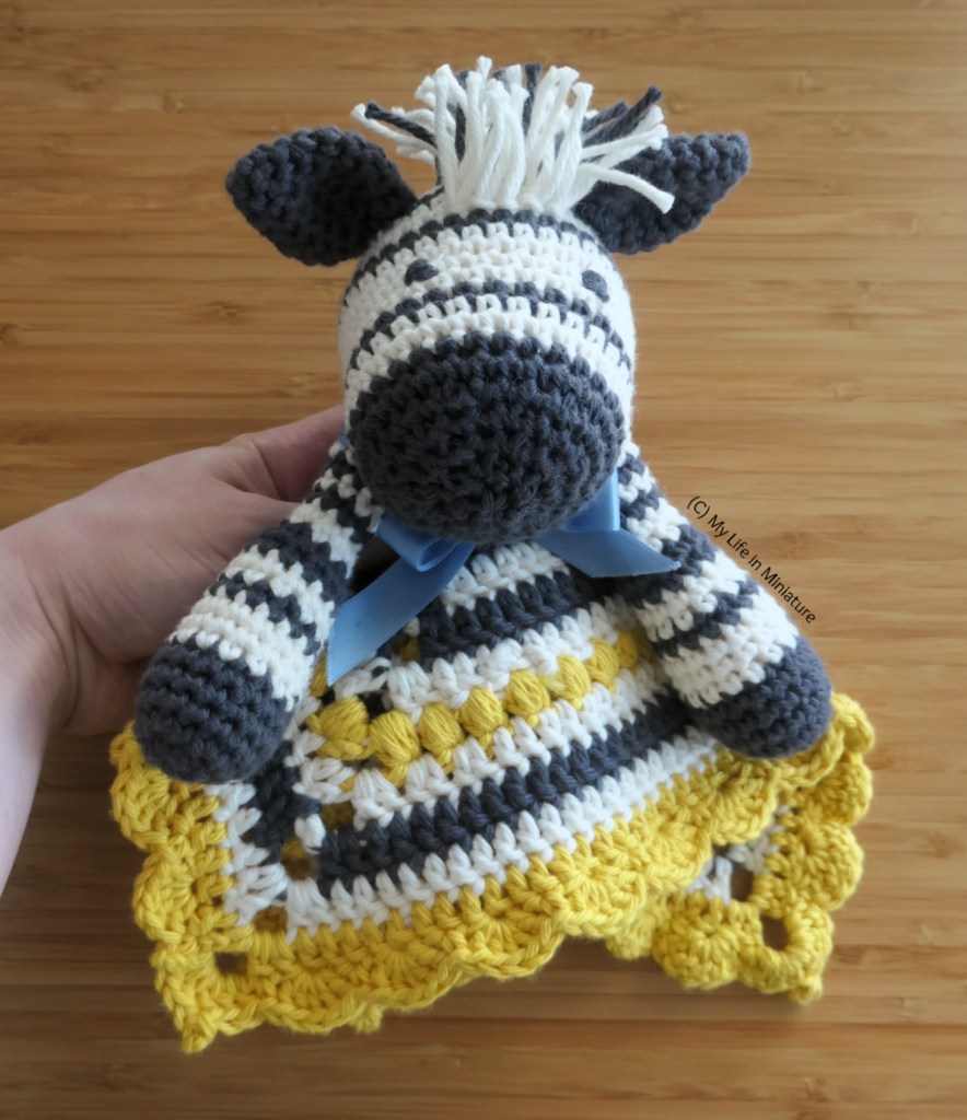 The author's hand holds the zebra toy in front of a wood background. The toy's face is facing the camera; it has black embroidered eyes.