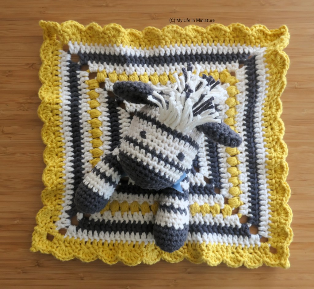 The zebra baby comforter is photographed from above on a wood surface. Its blanket body has been spread out flat beneath its head and arms. The black and white stripes, yellow scallops, and yellow puff stitches in the blanket are all visible. 