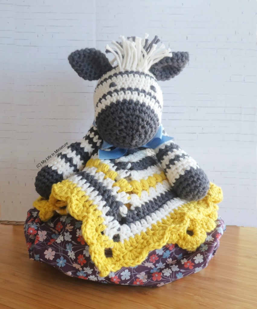 A crocheted zebra baby toy sits on a stuffed project bag, in front of a white brick background. It is a comforter, so its body is a blanket of black, white, and yellow stripes with a scalloped edge. It also has a blue bow around its neck.