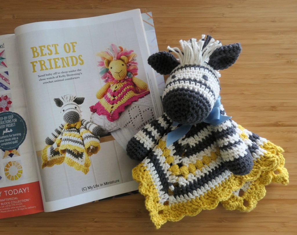 The zebra baby toy is laid next to an open magazine page, on a wood surface. The magazine is open to the cover page for the pattern for the toy. There are two similar crocheted comforters on the page: one the zebra, the other a yellow lion with pink accents. 
