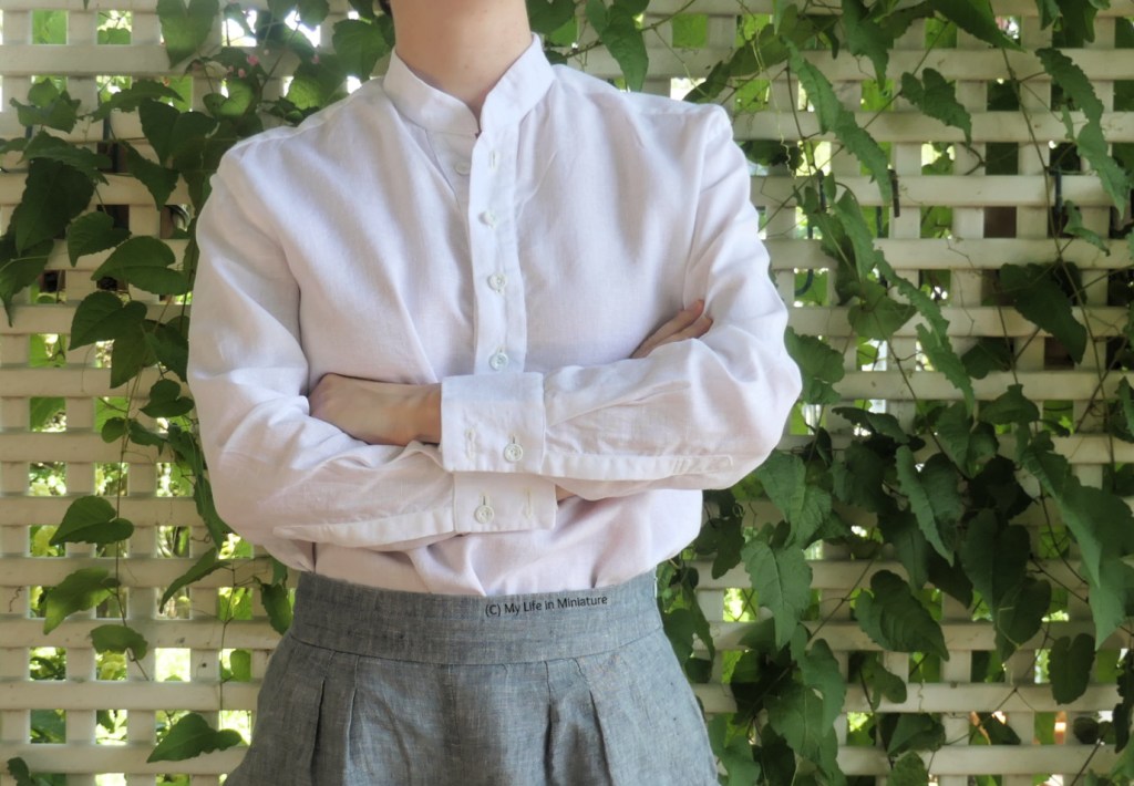The author stands in front of a lattice with a vine growing through it. She's photographed from neck to thighs, wearing a long-sleeved, collarless, white linen shirt tucked into pale grey pants. She faces the camera with her arms crossed. The shirt is tucked into her pants.