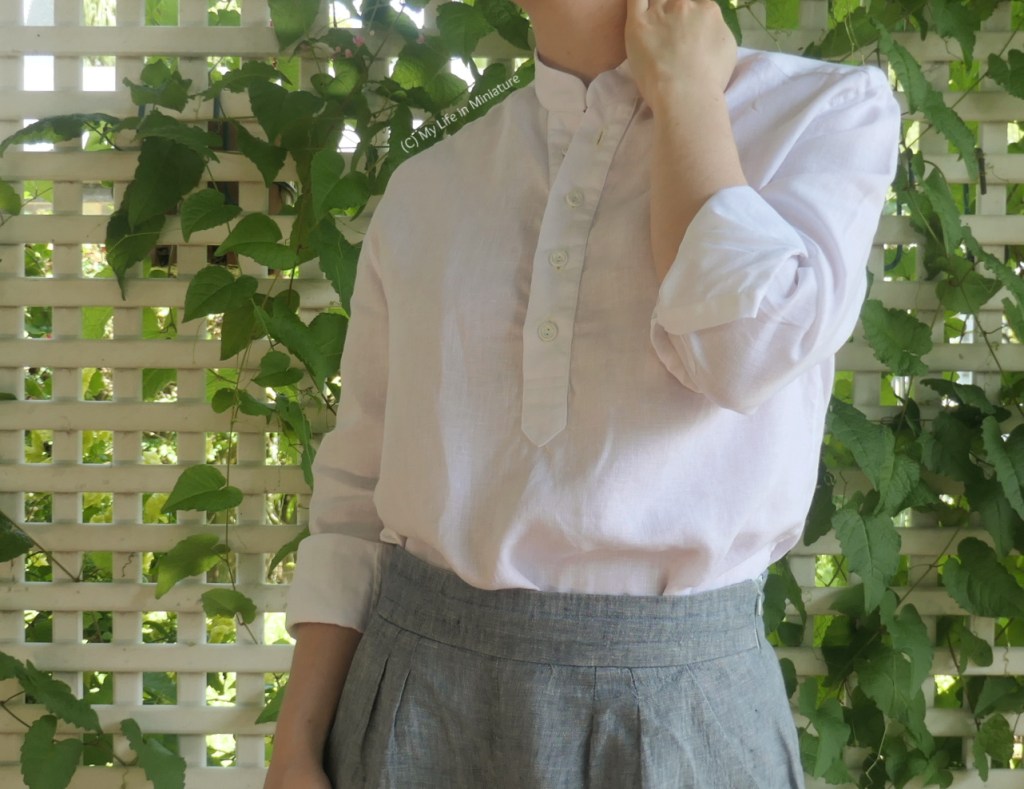 The author stands in front of a lattice with a vine growing through it. She's photographed from neck to thighs, wearing a long-sleeved, collarless, white linen shirt tucked into pale grey pants. The sleeves are rolled up, and the author has one hand up near her neck, with the other at her side. 