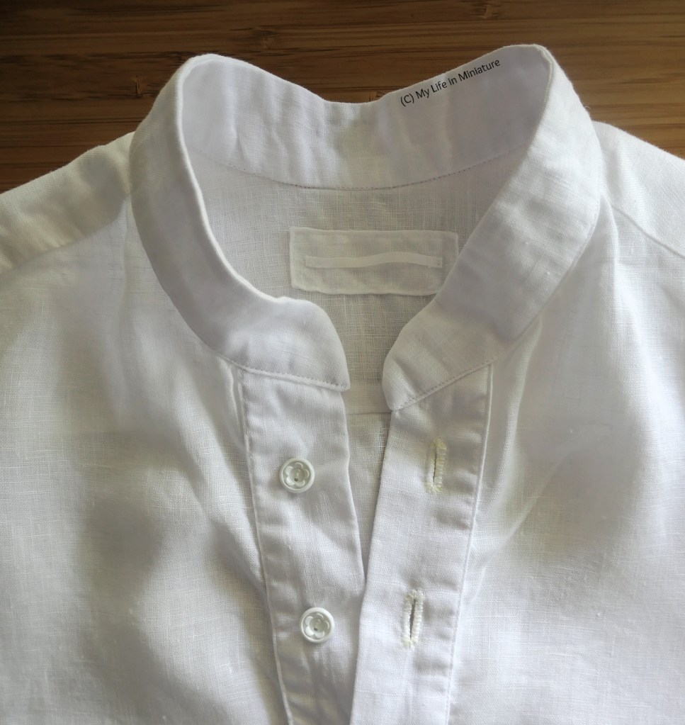 Closeup of the collar and centre front button placket, taken flat from above on a wood surface. The buttons on the button stand are small, white, and circular with little flowers inside. On the inside yoke, at the back fo the neck, is a hanging loop of white ribbon sewn to a small square of the linen.