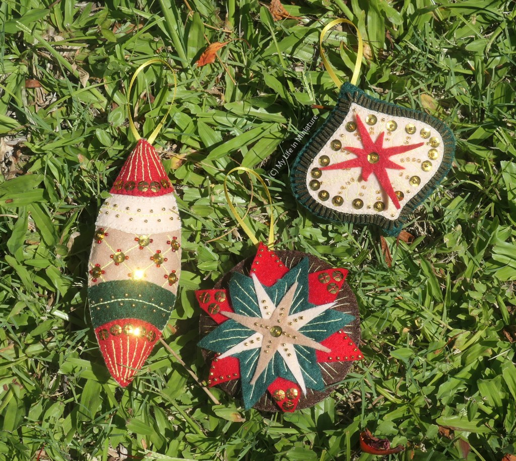 The three felt ornaments are laid flat on grass in the sun, photographed from above. Some of the sequins sewn onto the ornaments are sparkling in the sunlight. 