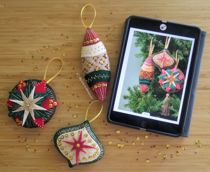 Three embroidered Christmas decorations are laid flat on a wood surface, next to a small iPad. Photographed from above, the decorations are heavily embroidered and beaded. The iPad has an image on the screen of similar decorations, used to guide a tutorial from a magazine.