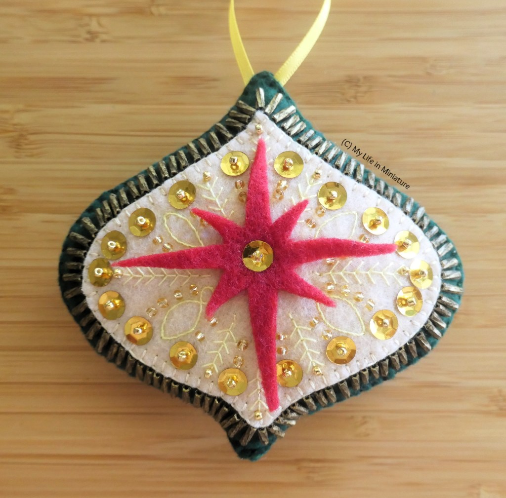 The third ornament is photographed up close on a wood surface. It is oval-ish, tapering to points at the top and bottom, and is dark green. In the centre is an eight-pointed red star, with a sequin sewn to the centre. Behind it is an oval-ish shape in cream, heavily embroidered with stem-and-leaf shapes, and heavily sequinned and beaded. The gap between the cream shape and the edge of the green shape is beaded with small, tubular, dirty gold beads, radiating out from the centre. At the top is a yellow loop of ribbon.