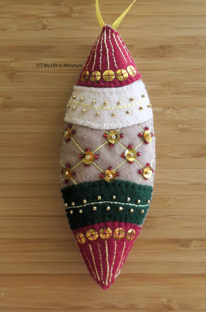 One of the ornaments, photographed up close. It is a long oval shape, with pointed ends. It's separated into five sections; the top and bottom-most sections are red, with vertical embroidered lines and gold sequins. The middle section is light brown, with an embroidered pattern like a plush leather seat, and sequins and beads sewn on where the lines cross. The final two sections are bands with an embroidered line across the centre, and gold seed beads along each side. One band is cream, the other is dark green. The ornament has a yellow loop of ribbon at the top to hang it.