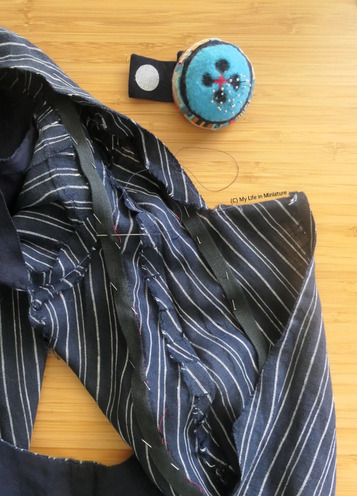 The insides of a navy striped waistcoat are laid flat on a wood surface. Strips of black cotton tape are pinned to the inside, and a needle is in the process of stitching them down. Above the waistcoat is a pincushion.