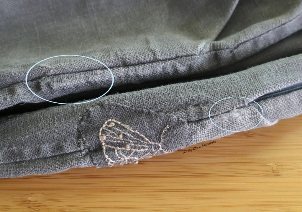 Close-up of the side seams of a pair of dark grey linen pants, laid on a wood surface. Two areas of wear at the seams are circled with light blue graphics. 