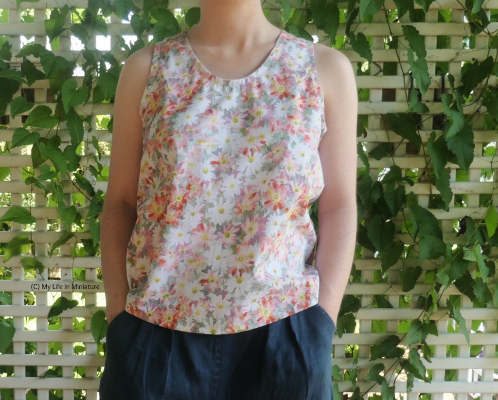 The author stands in front of a lattice with a vine growing through it. She's photographed from neck to thighs. She is wearing a sleeveless top with navy pants, and her hands are in her pant pockets. The top's fabric has pink, red, purple, and white flowers on it, in very washed-out, pastel hues. The neckline is curved. 
