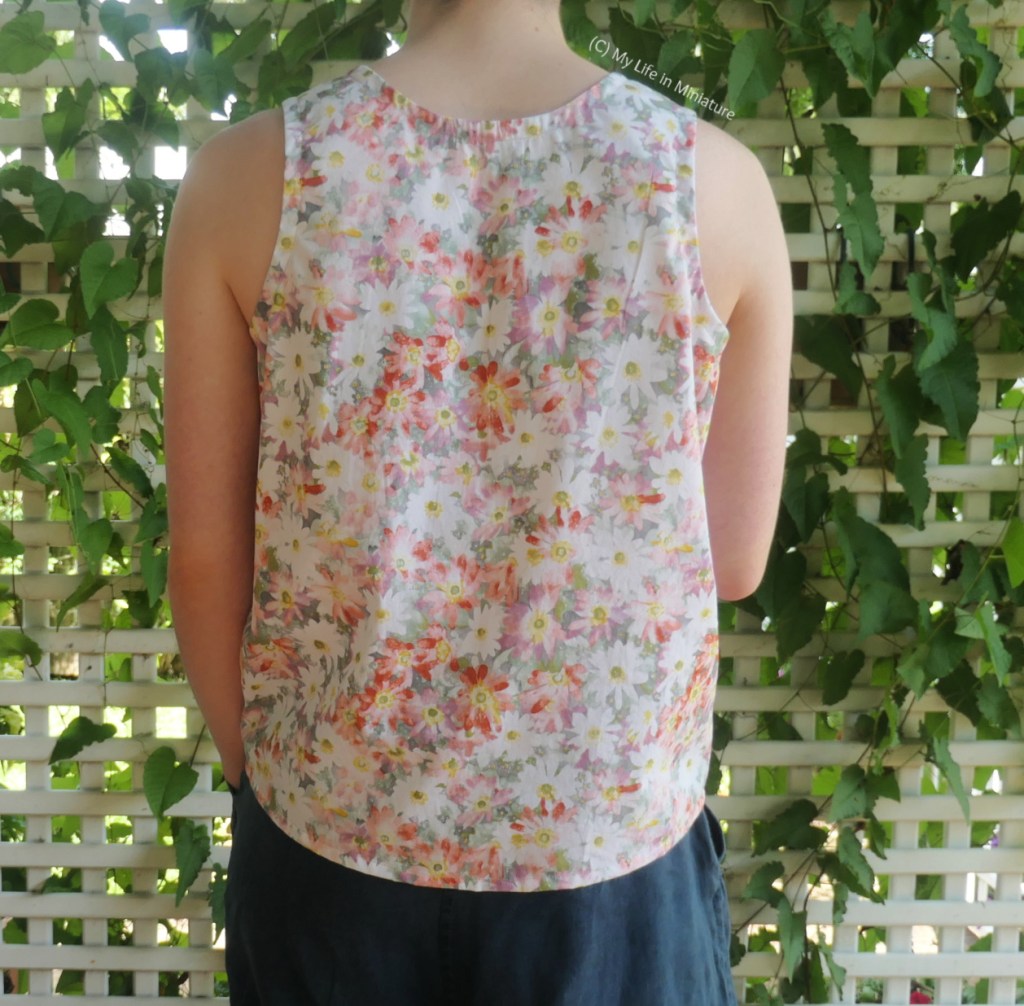 The author stands in front of a lattice with a vine growing through it. She's photographed from neck to thighs. She has her back to the camera, so the back of her sleeveless top is visible. The back neckline is also scooped a little, and the back hem is longer than the front hem. She still wears her navy pants.