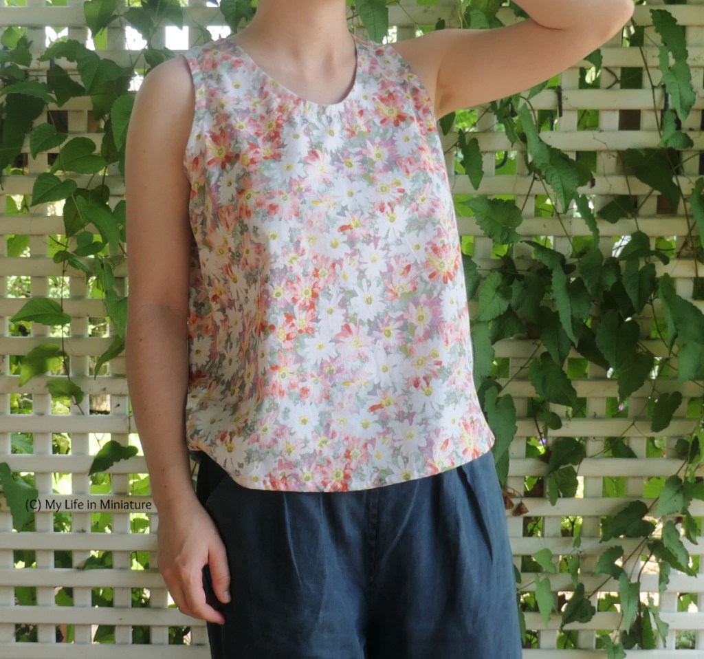 The author stands in front of a lattice with a vine growing through it. She's photographed from neck to thighs. Still wearing the sleeveless top and navy pants, she has one hand hanging at her side and the other raised out of frame. She's slightly angled to the right of frame.