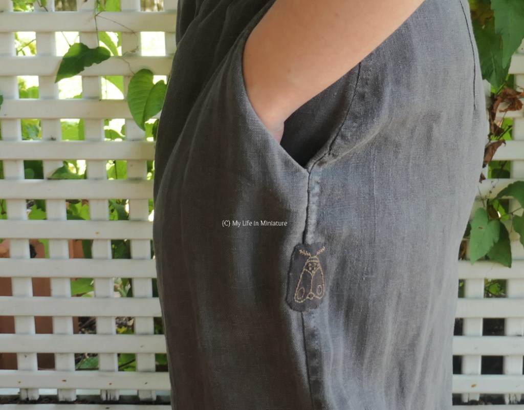The author stands in front of a lattice with a vine growing up it. In frame is a closeup of one of the pockets of the dark grey pants as the author wears them. At the seam, roughly over where the wear was earlier, is an embroidered moth patch. The author's hand is in the nearby pocket.