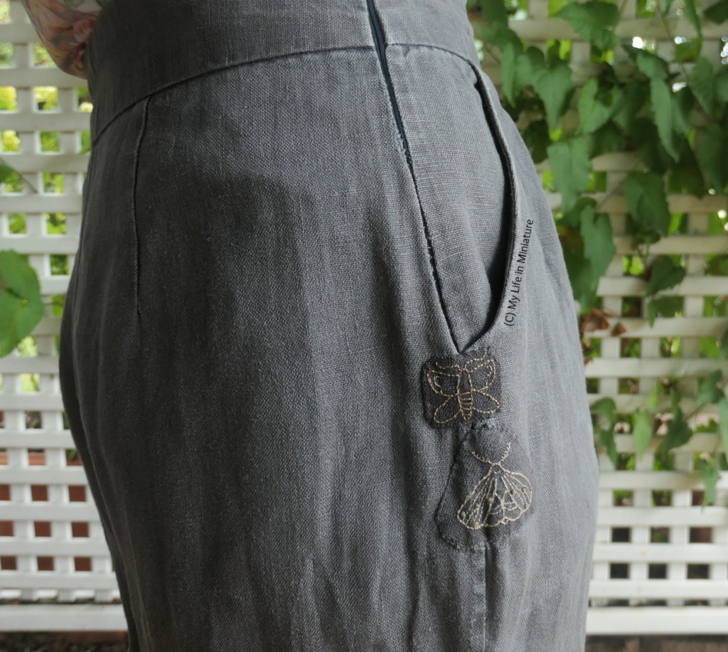 The author stands in front of a lattice with a vine growing up it. In frame is a closeup the other pocket of the dark grey pants as the author wears them. Above the previous moth patch is another, smaller, embroidered mtoh patch. 
