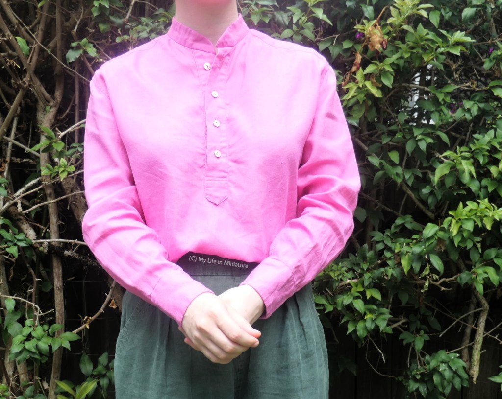 The author wears the pink button-up shirt tucked into green pants. She's buttoning the cuff of one sleeve, with her torso facing the camera. The shirt has no collar, and the button placket finishes about midway down with a point. 