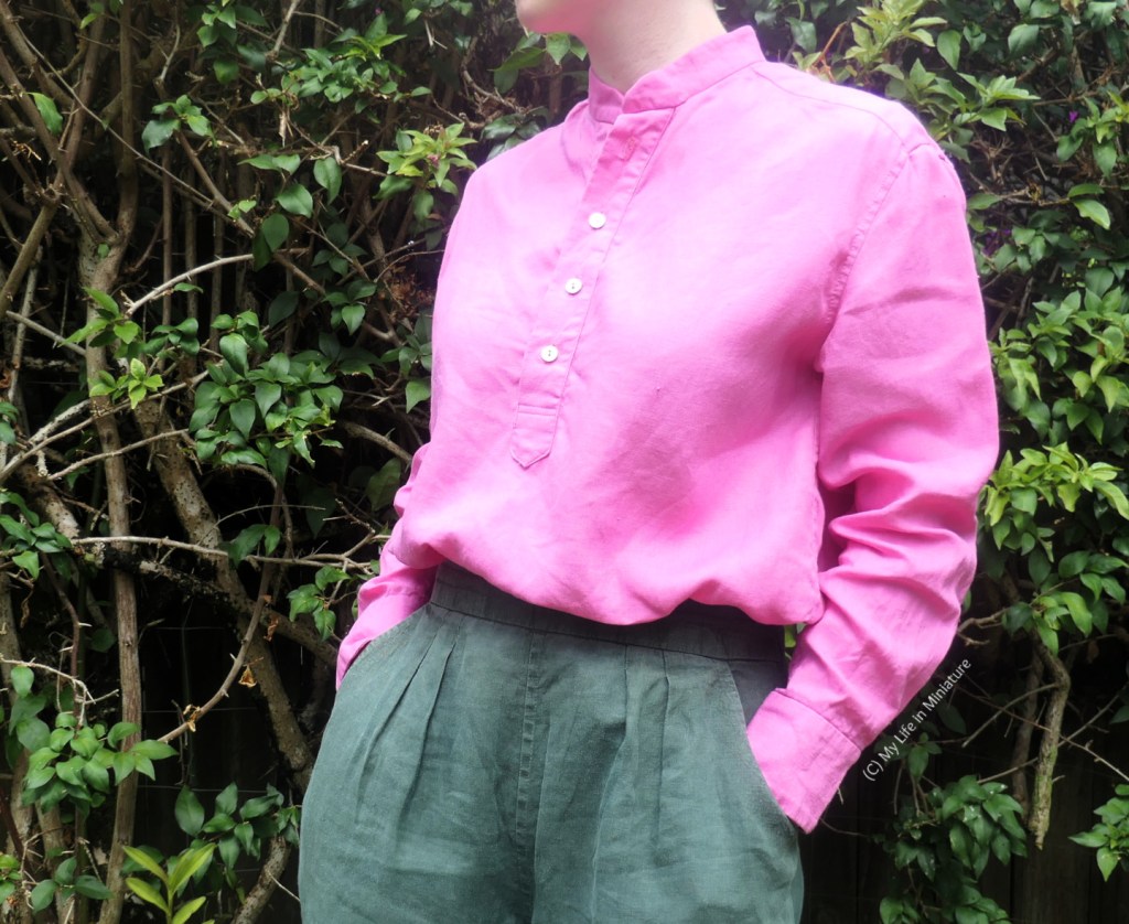 The author stands in front of a hedge throughout, photographed from neck to thighs. She's wearing a bright pink button-up shirt with dark green pants. The shirt is tucked into her pants, and her body is at a three-quarters angle to the camera. Her hands are tucked into her pant pockets.