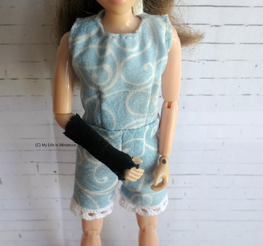 Close-up of Sarah's right arm as she's holding it in front of her torso. A black tube of knit fabric encases the arm from her elbow to her knuckles. She's wearing a pale blue playsuit, and is standing against a white brick background.
