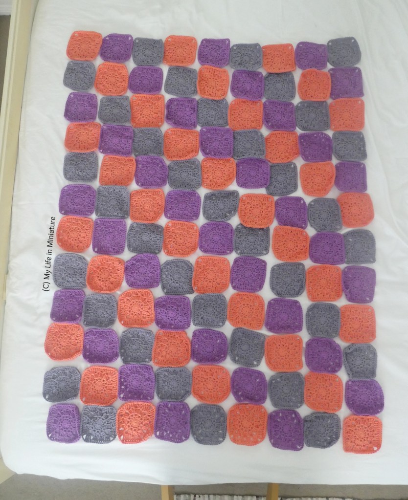 108 crochet squares are laid out in a rectangle on a bed, made with a white sheet. They are arranged in diagonal lines of peach, purple, and grey.