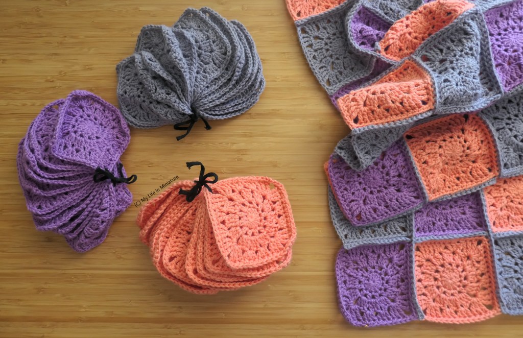 Three stacks of crochet squares, tied together with string, are arranged in a rough C shape on a wood background. Next to them is a small crocheted blanket, made of more of the same squares. It's folded prettily, and extends further out of frame.