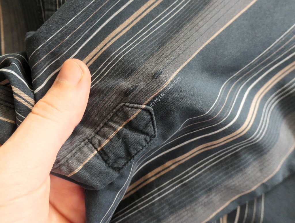Close-up of the sleeve of a black shirt with irregular tan and white stripes. Near the base of the sleeve placket are two small rips in the fabric. The author's hand is holding the rips closer to the camera.