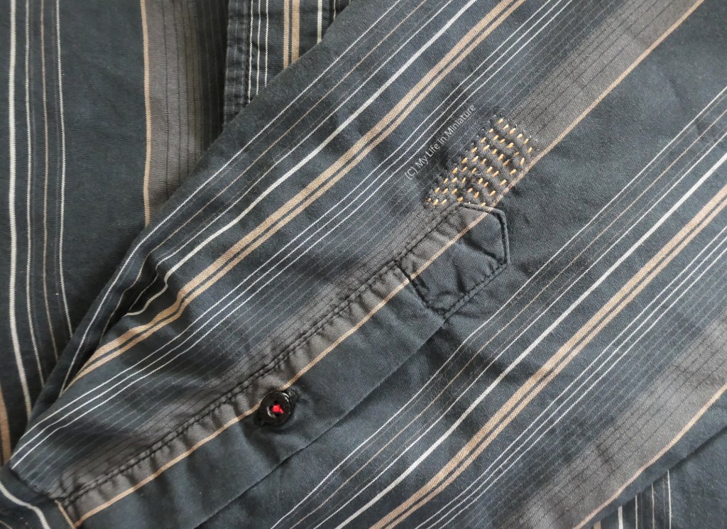 The outside of the mended sleeve of the black-white-tan-striped shirt. Near the base of the sleeve placket is a small rectangle of fairly consistent running stitches, done in yellow thread.