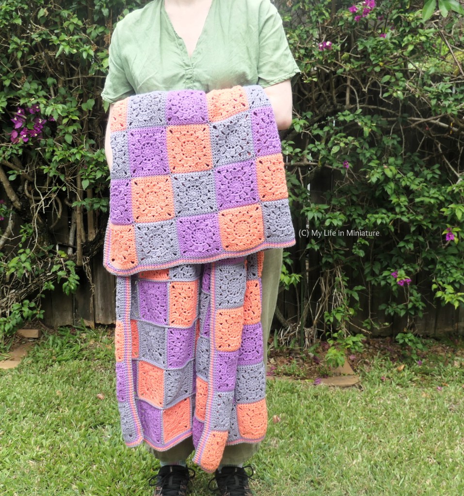 The author stands in front of a hedge, with the crocheted blanket loosely tossed over one forearm. She's photographed from shoulders to ground, and is wearing green linen top to toe. The blanket's border is in the colours used in the blanket itself, and is quite simple.