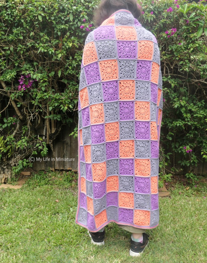 The author stands in front of a hedge, facing away from the camera. She has the crocheted blanket wrapped around her shoulders, and it covers the majority of her body. (The back of her head and her heels are visible at each end of the blanket.)