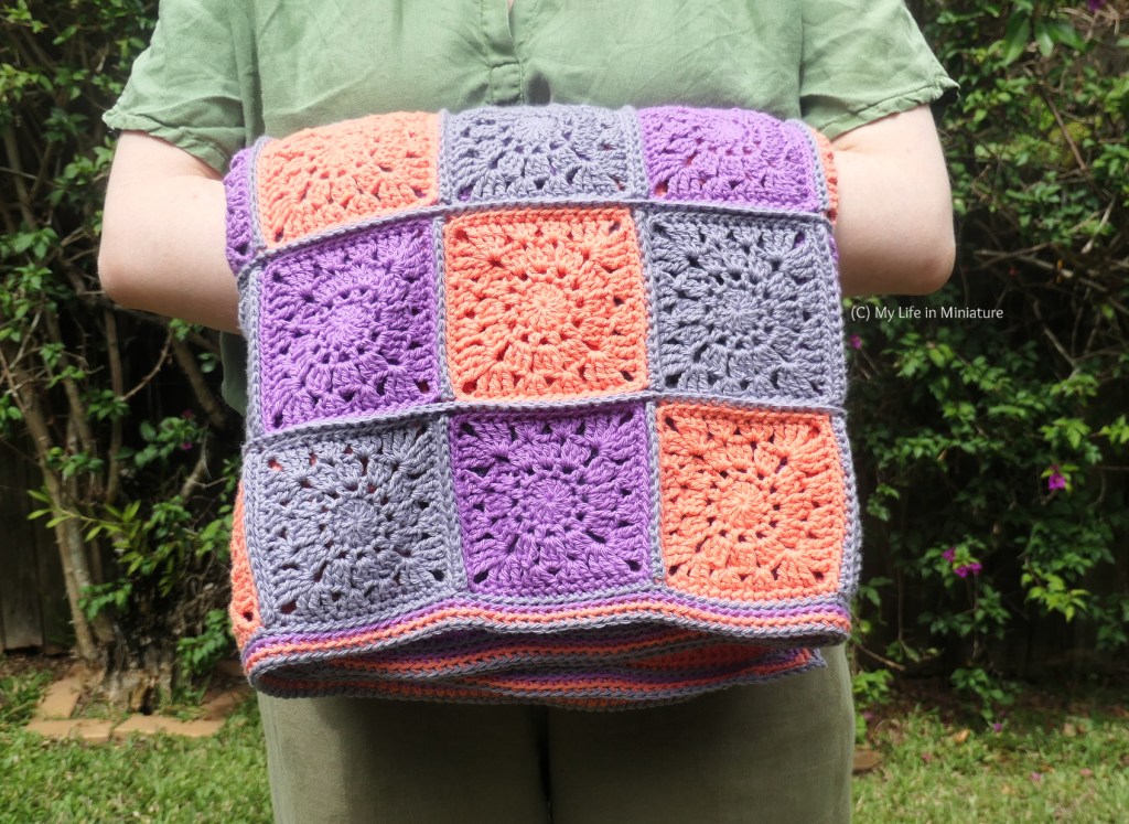 Close-up of the blanket, neatly folded in the author's arms. The border is four rows of plain crochet, done into the back loop so there is a ridge between each colour. The squares are a little bit lacy, and are connected with the grey yarn.