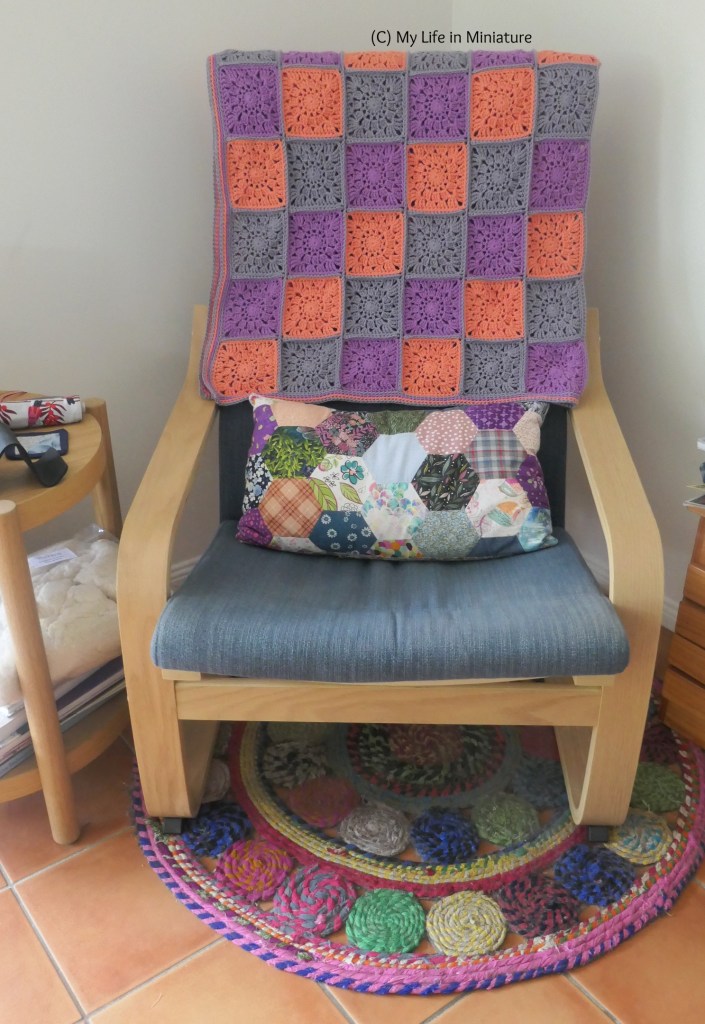 The crocheted blanket is folded neatly and draped over the back of an armchair in a corner inside the author's house. The armshair has navy upholstery, and is made of wood. Also on the armchair is a rectangular cushion, longer than it is wide, with a hexagonal patchworked cover. Just visible to the left is a small table, with a small mess on it.