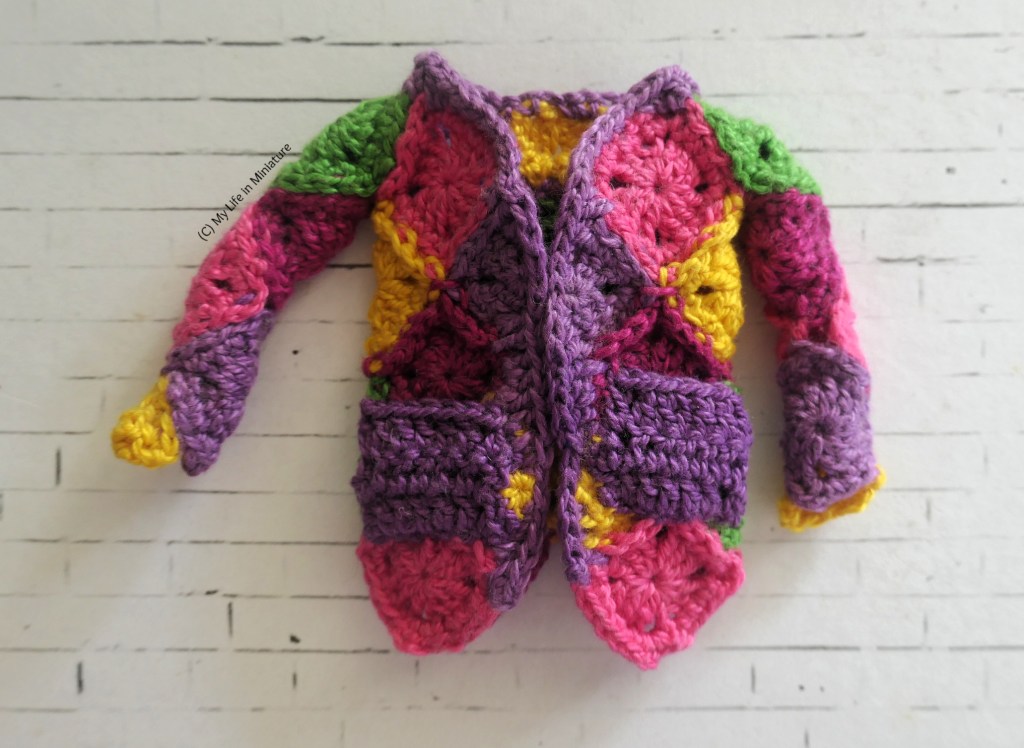 A miniature crochet cardigan is laid flat on a white brick background. It is made of crocheted squares in a variety of bright colours, with purple pockets and trim. It is a little bit janky.