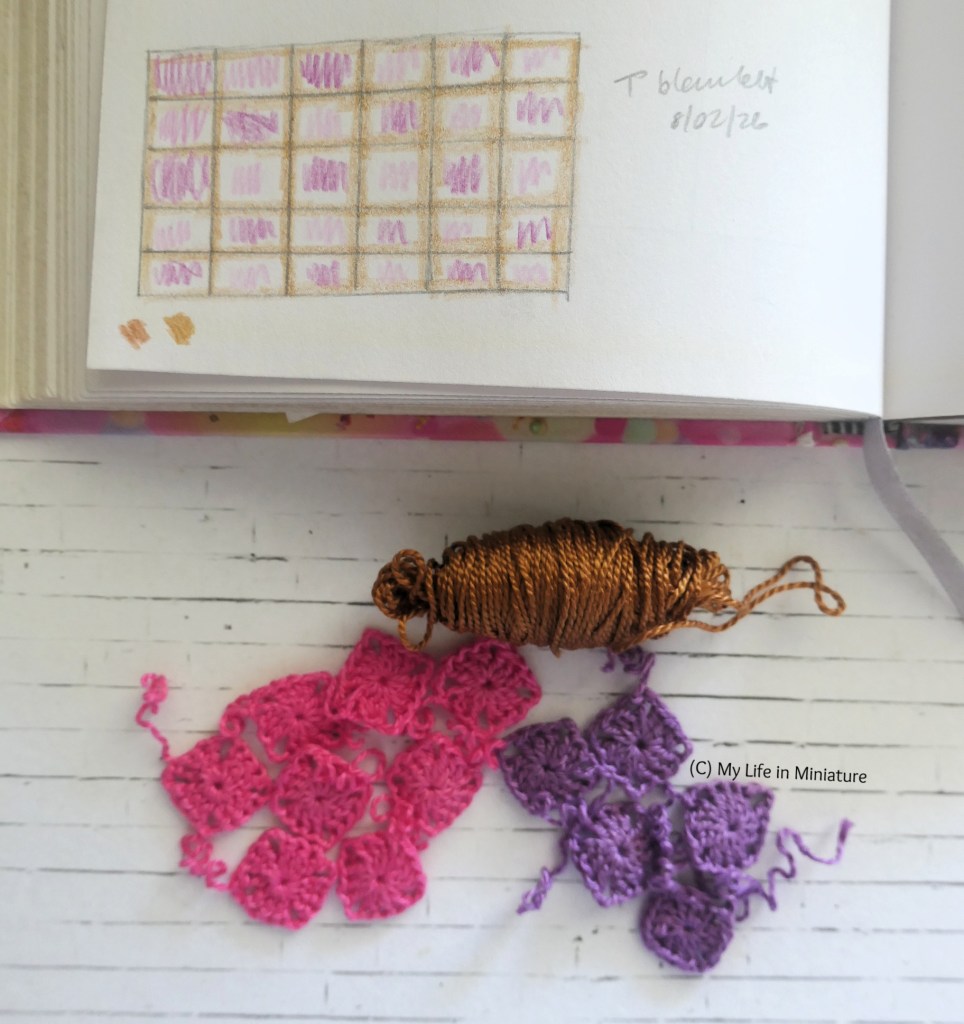 An open sketchbook and a small collection of crocheted squares are on a white brick background. The sketchbook is open to a sketch of a blanket. It is six by five squares, with each square coloured in pattern and the borders outlined. Below the sketchbook are two piles of crochet squares, one pink and one purple. There is also a small skein of copper yarn with the squares.