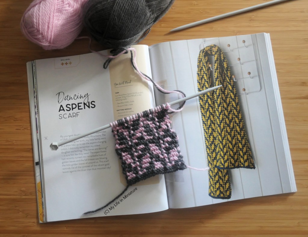 The library book from earlier is open to a page titled "Dancing Aspens Scarf", with an image of a leafy scarf beside the title. On the pages is a knitted sample of the pattern, still on the needles. The sample is knitted in pink and dark grey, and the yarn balls sit above the book. One half of the sample has the leaves in pink and the background grey; the other half of the sample reverses the colours.