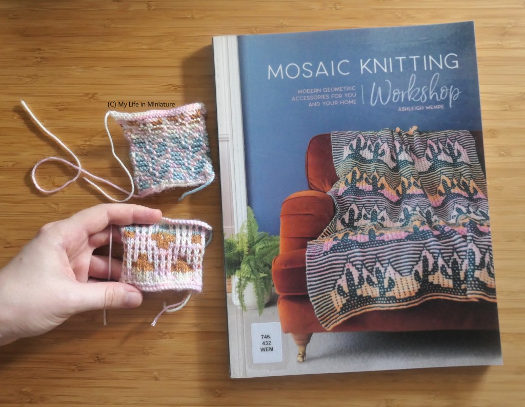 A book and two knitted swatches are photographed from above on a wood surface. The book is "Mosaic Knitting Workshop" by Ashleigh Wempe, with a library sticker in the bottom corner. To the left of it are the two knitted swatches. The author's hand holds one flat. 